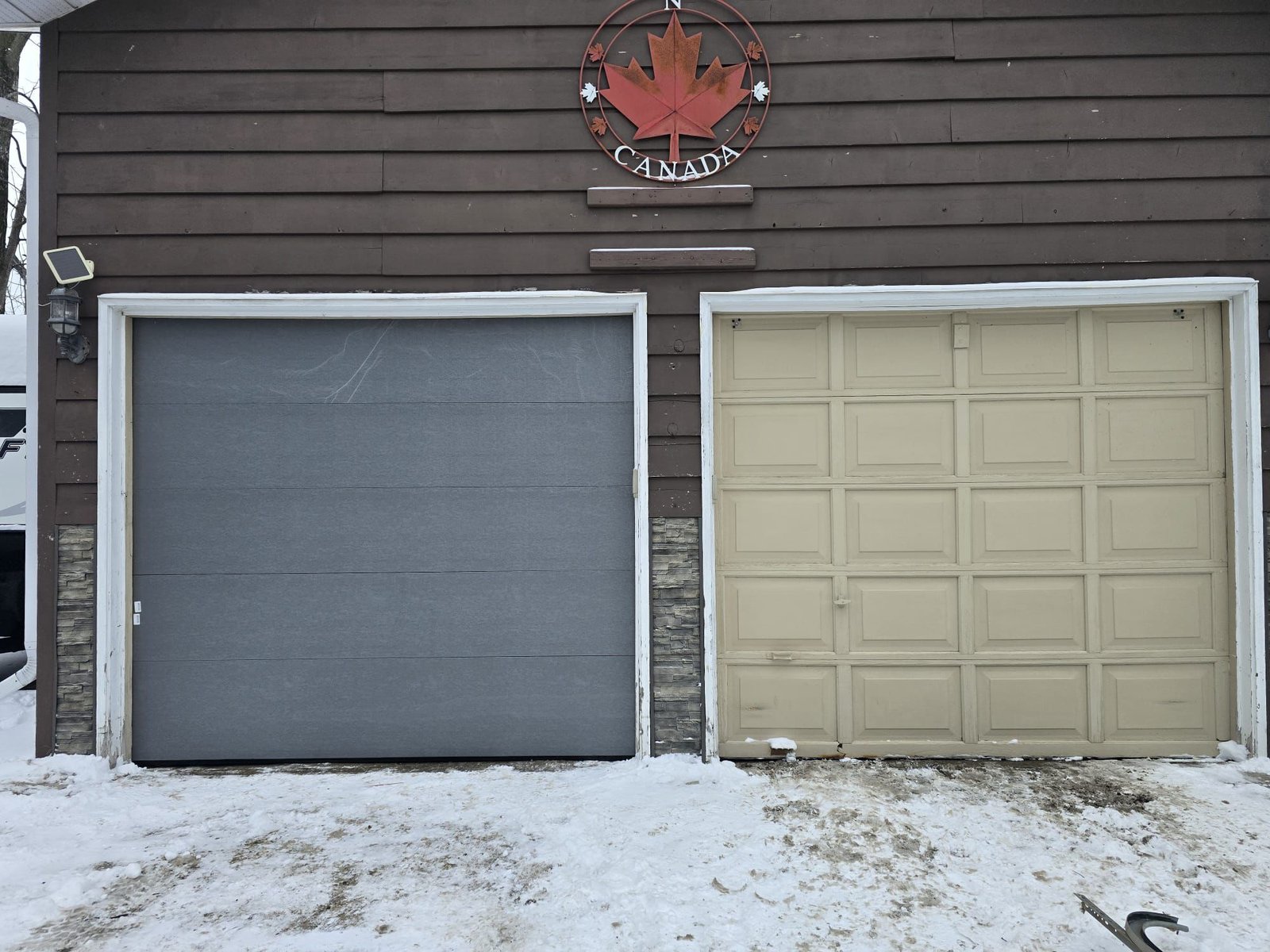 A newly installed grey garage door beside an older beige raised-panel door on a wood-sided home